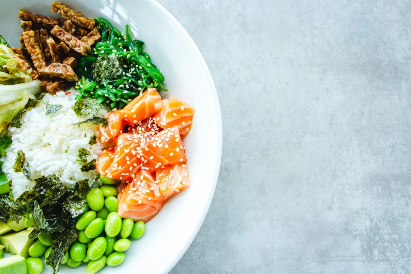 Fresh salmon poke bowl with rice, avocado, edamame, seaweed, and sesame seeds in a white bowl on grey background