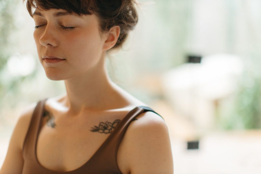 Woman meditating with eyes closed indoors, showing lotus tattoos on shoulders
